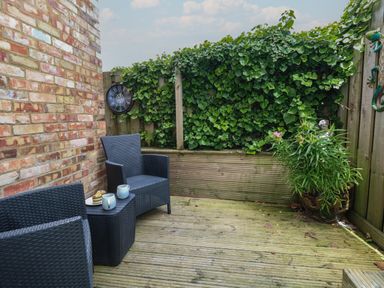 A small outdoor patio with two black wicker chairs a small table with mugs and food a brick wall green ivy on a wooden fence and a flower pot at Flat 1 - Sandbanks in Bournemouth