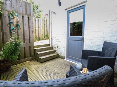 A small outdoor patio with wooden decking chairs a table with snacks a potted plant wooden fencing and a blue door at Flat 1 - Sandbanks in Bournemouth
