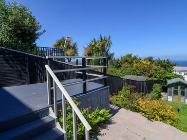 An outdoor garden area with stairs leading to a deck and plants with trees and a shed in the background at Combe View in Ilfracombe