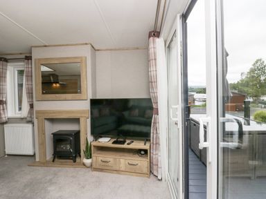 A living room with a television on a wooden stand, an electric stove in a fireplace, a mirror above, curtains on windows, and a sliding glass door leading to an outdoor seating area at Mon in Troutbeck Bridge