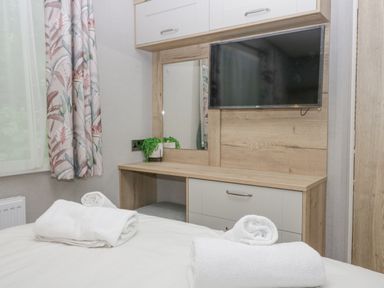 A bedroom with a bed with rolled white towels a wooden dresser a mirror a wall-mounted television and floral curtains at Mon in Troutbeck Bridge
