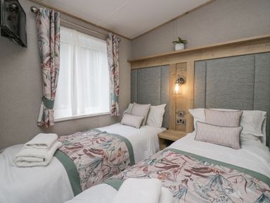 A bedroom with two single beds each with floral bed runners and cushions a window with floral curtains and a wall lamp above a wooden headboard at Mon in Troutbeck Bridge