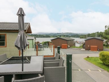 An outdoor seating area with a glass table and wicker chairs on a deck near wooden cabins by a lake with hills in the background at Mon in Troutbeck Bridge