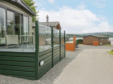 An outdoor patio with glass railing and wicker chairs next to a paved road and wooden cabins with a lake and hills in the background at Mon in Troutbeck Bridge