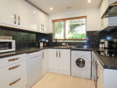 A kitchen with white cabinets black countertops a washing machine dishwasher microwave and a window with blinds at Tincroft in Porthtowan