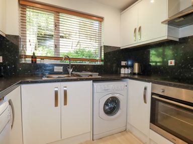 A kitchen with white cabinets a washing machine an oven a sink and a window with blinds at Tincroft in Porthtowan
