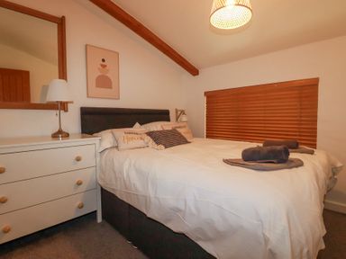 A bedroom with a double bed, white dresser with lamp, wooden framed mirror, and window with wooden blinds at Tincroft in Porthtowan