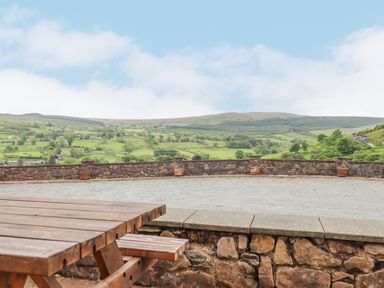 A wooden picnic table on a stone patio overlooking a stone wall and green hills at Llys-y-Wennol in Penmachno