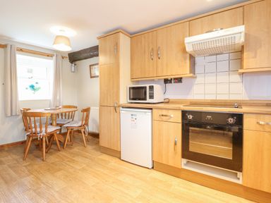 A kitchen with wooden cabinets a microwave oven a small refrigerator a dining table with four chairs and a window at Llys-y-Wennol in Penmachno