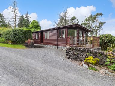 A gravel driveway leading to a wooden lodge with a stone base and a small deck with outdoor furniture surrounded by trees and shrubs at Lodge 3 At Pound Farm in Crook near Kendal