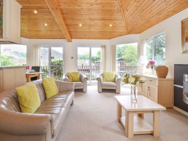 A living room with beige sofas and yellow pillows a wooden coffee table with flowers a wooden ceiling and large windows at Lodge 3 At Pound Farm in Crook near Kendal