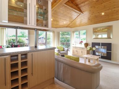 A living room with a wooden ceiling a sofa and armchair a fireplace a mirror a cabinet with flowers and glass shelves with glasses at Lodge 3 At Pound Farm in Crook near Kendal