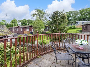 An outdoor patio with a table and chairs and wooden railing overlooking garden and wooden lodges at Lodge 3 At Pound Farm in Crook near Kendal