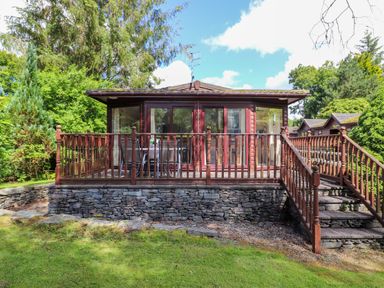 A wooden lodge with a raised stone foundation and a wooden deck surrounded by trees at Lodge 7 At Pound Farm in Crook near Kendal