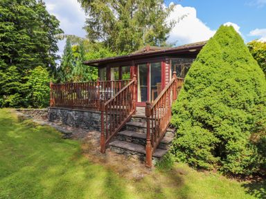 A wooden deck with railing and stairs leading to a cabin surrounded by trees and shrubs at Lodge 7 At Pound Farm in Crook near Kendal