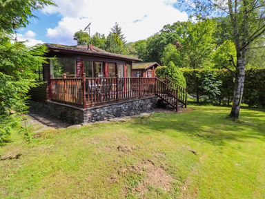 A wooden lodge with a deck and railing surrounded by grass trees and bushes at Lodge 7 At Pound Farm in Crook near Kendal