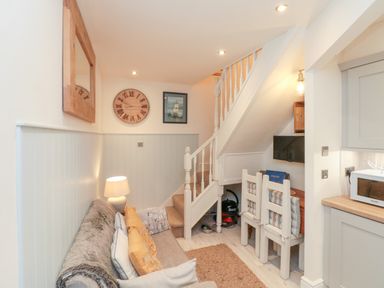 A small living area with a sofa pillows lamp wall clock and framed picture next to a staircase with two chairs and a television under the stairs at Little Gem in Whitby