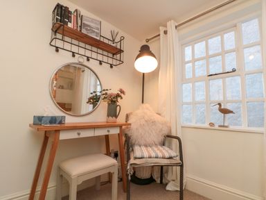 A corner with a wooden desk and stool a round mirror on the wall a chair with cushions a window and a floor lamp at Little Gem in Whitby