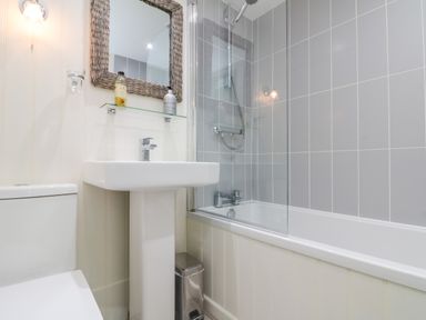 A bathroom with a toilet, pedestal sink, bathtub with shower, mirror, and wall shelf at Little Gem in Whitby