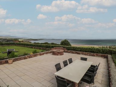 An outdoor patio with a dining table and chairs next to a fire pit and a grassy area with a barbecue overlooking the sea at Cois Leice Cushlecka near Mulranny County Mayo