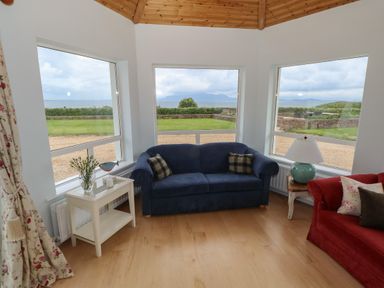 A sitting area with blue and red sofas a white side table with decor and large windows showing a garden and hills at Cois Leice in Cushlecka near Mulranny County Mayo