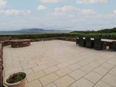 An outdoor patio with a fire pit built into a stone bench and a long table with chairs overlooking a body of water at Cois Leice Cushlecka near Mulranny County Mayo