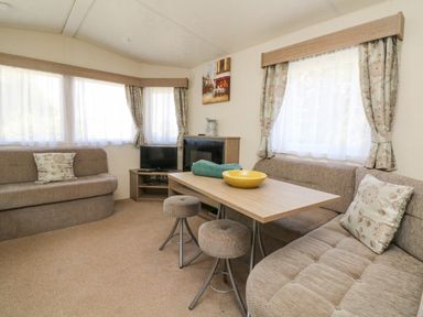 A living area with sofas a wooden table two stools a television and windows with curtains at 23 Manleigh Park in Combe Martin