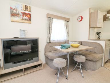 A dining area with a corner sofa seating a table and two stools next to a fireplace and a window with curtains at 23 Manleigh Park in Combe Martin