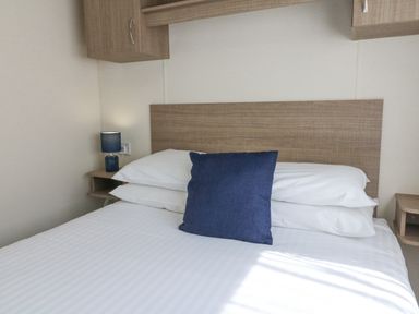 A bed with a blue pillow and white sheets next to a bedside table with a lamp at 23 Manleigh Park in Combe Martin