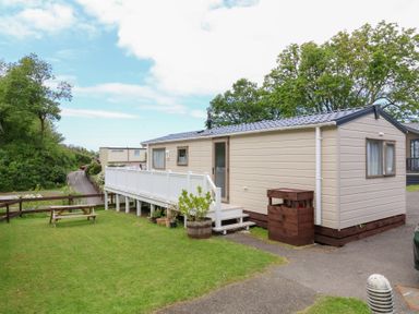 A beige mobile home with white railings and stairs on a lawn with a wooden picnic table and trees at 23 Manleigh Park in Combe Martin