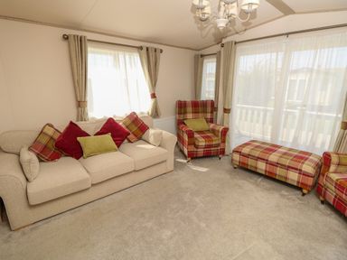 A living room with a beige sofa with cushions a plaid armchair and ottoman with curtains and windows at The Boathouse in East Wittering