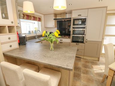 A kitchen with a countertop island with sunflowers in a vase two chairs built in oven microwave and a window at The Boathouse in East Wittering