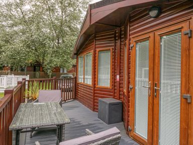 An outdoor wooden deck with a table, two chairs, a storage box, and a wooden lodge with windows and glass doors at Don's Lodge in Grasmere 3 White Cross Bay near Windermere