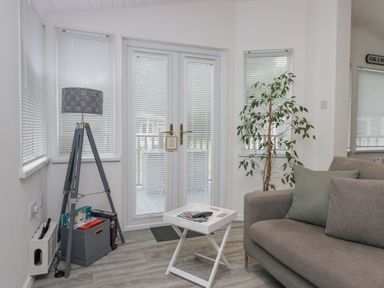 A living room with a gray sofa a white folding tray table with remote controls a tall lamp with a basket at the base and glass doors with blinds at Don's Lodge in Grasmere 3 White Cross Bay near Windermere