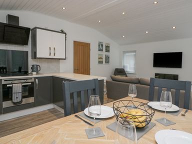 A kitchen and dining area with a table set with plates and glasses overlooking a living room with a sofa and wall-mounted TV at Don's Lodge in Grasmere 3 White Cross Bay near Windermere