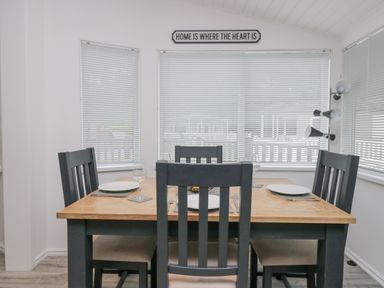 A dining area with a wooden table, four black chairs, plates and glasses set and a sign on the wall reading home is where the heart is at Don's Lodge in Grasmere 3 White Cross Bay near Windermere