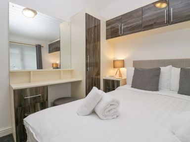 A bedroom with a bed, rolled towels on the bed, bedside table with a lamp, a vanity desk with a mirror and drawers at Don's Lodge Grasmere 3 White Cross Bay near Windermere