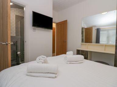 A bedroom with a bed, folded towels, a wall-mounted television, a vanity desk with a mirror and two open doors at Don's Lodge Grasmere 3, White Cross Bay near Windermere