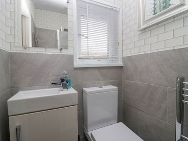 A bathroom with a sink cabinet, a toilet, a window with blinds, a wall mirror, and a framed picture at Don's Lodge in Grasmere 3 White Cross Bay near Windermere