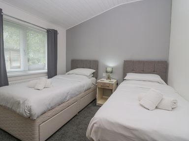 A bedroom with two single beds with white bedding a nightstand with a lamp and window with blinds and curtains at Don's Lodge Grasmere 3 White Cross Bay near Windermere