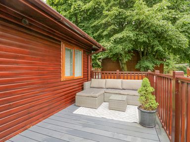 An outdoor seating area with a sectional sofa and small table on a deck with wooden walls and railing with a potted plant at Don's Lodge in Grasmere 3 White Cross Bay near Windermere
