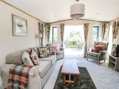 A living room with a sofa, armchairs, coffee table, patterned curtains, and a view outside at Saorsa in St Fillans