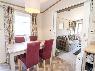 A dining area with a wooden table and four red chairs next to a living room with a gray sofa and floral cushions at Saorsa in St Fillans