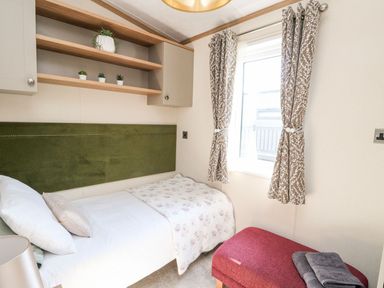 A bedroom with a single bed white bedding and a green headboard with shelves and small plants above a window with patterned curtains and a red bench with folded towels at Saorsa in St Fillans