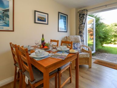 A dining area with a wooden table set for four with bowls glasses and a fruit bowl next to a wicker chair by an open glass door to a garden at 74 Reap Lane Southwell near Weston