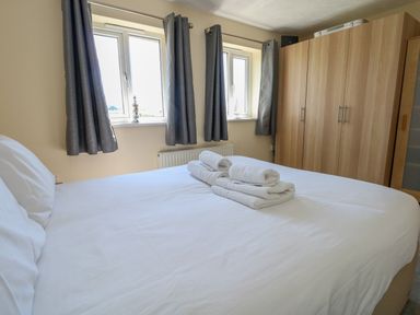 A bedroom with a white bed and folded towels on it two windows with grey curtains and wooden wardrobes at 74 Reap Lane Southwell near Weston