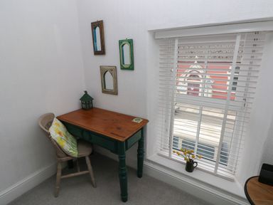 A small wooden desk with a chair and cushion next to a window with blinds and a small vase with flowers on the windowsill at Bryn Siriol in Narberth