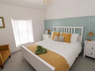 A bedroom with a white bedframe a bed with yellow and floral pillows green towels on the bed white bedside tables with orange lamps a mustard armchair and a window with white blinds at Bryn Siriol in Narberth