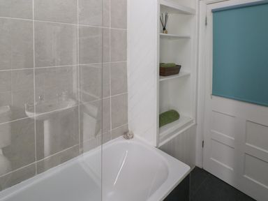 A bathroom with a bathtub a glass shower screen built in shelves with towels and a door with a blue blind at Bryn Siriol in Narberth