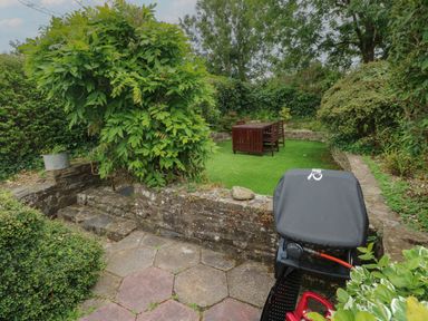 A garden patio with stone steps a covered barbecue and a wooden table with chairs on artificial grass at Bryn Siriol in Narberth